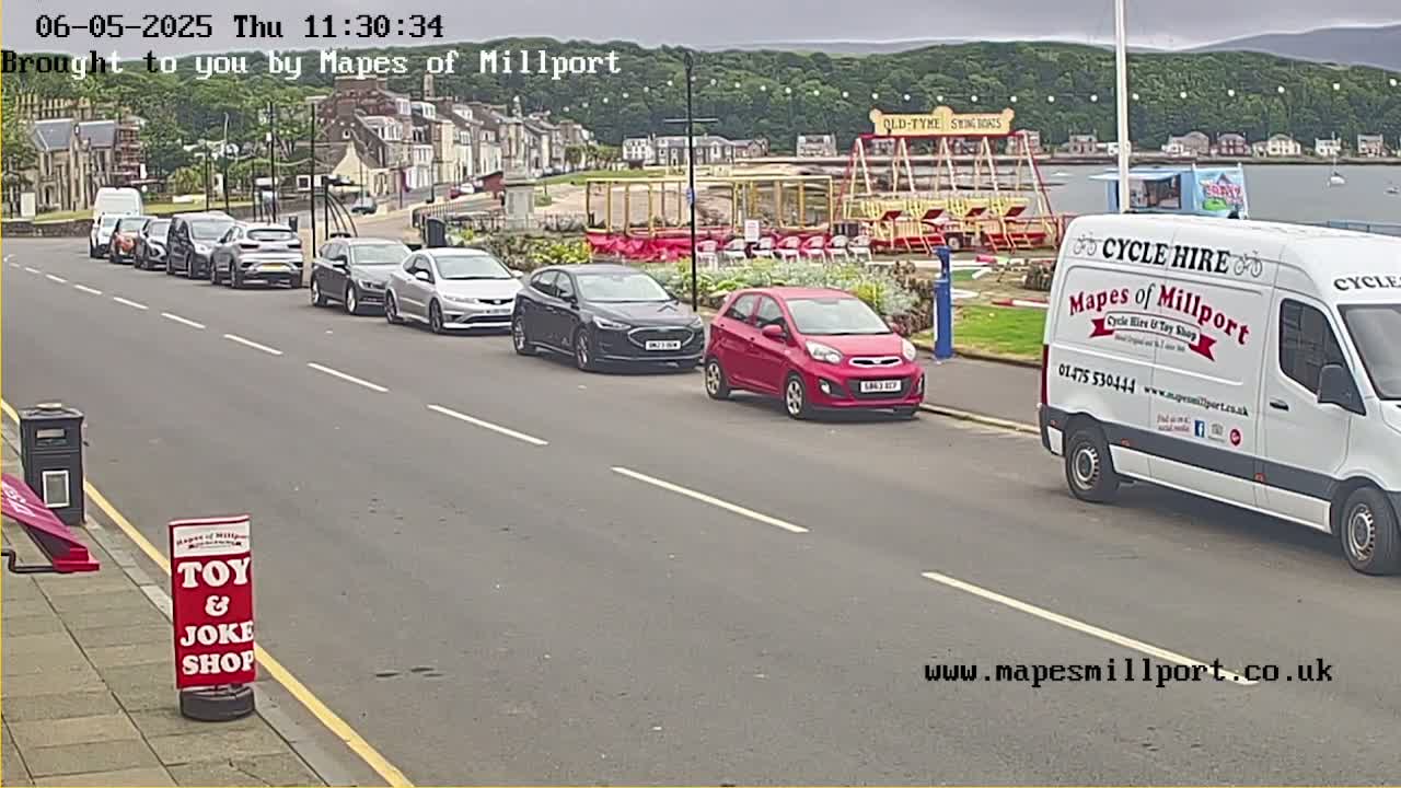 Several cars are parked along a seaside street on an overcast day, near a carousel and a white van.