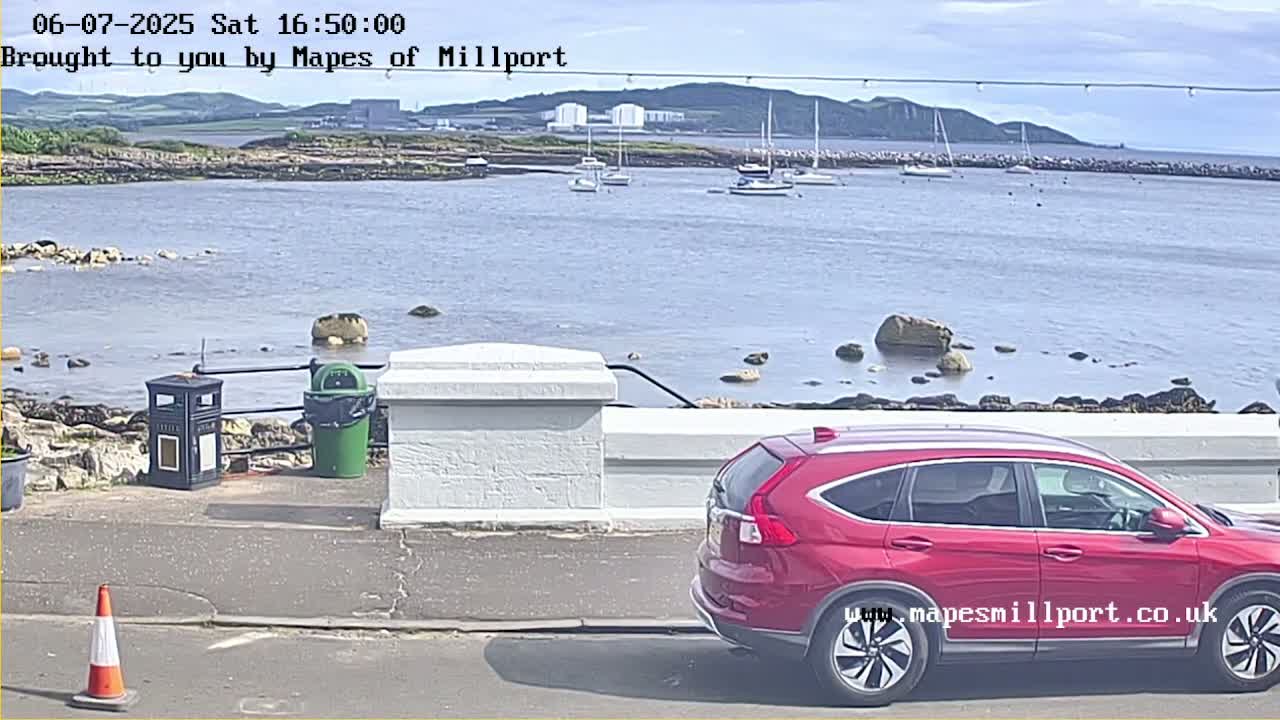 A red SUV is parked near a low white wall overlooking a calm body of water with several sailboats and a distant shoreline on a sunny day.