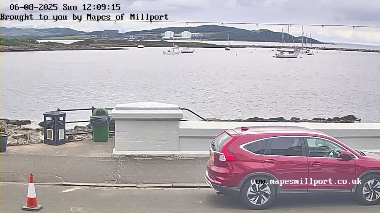 A red SUV is parked near a low white wall overlooking a calm body of water with several sailboats and a distant shoreline under overcast skies.
