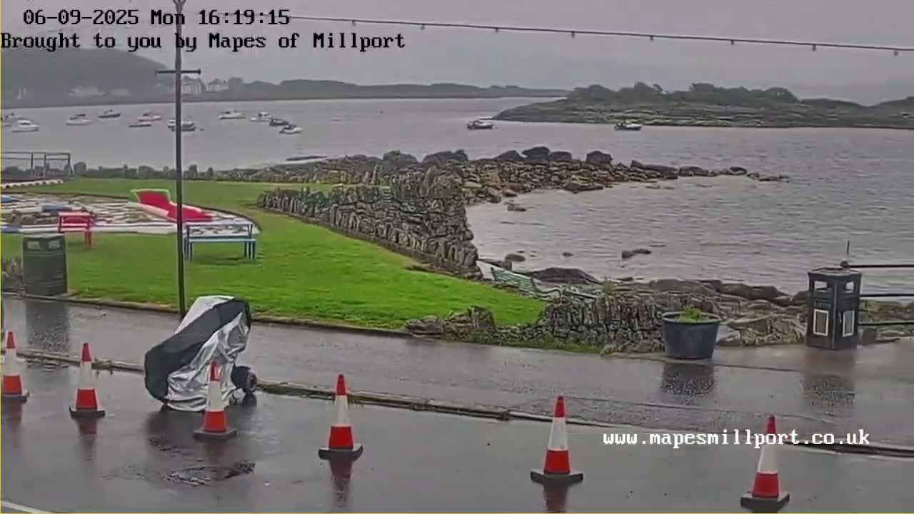 A wet, overcast day at a coastal location shows a rocky shoreline with several boats in the distance, a grassy area with playground equipment, and a vehicle covered by a tarp parked near traffic cones on a wet road.