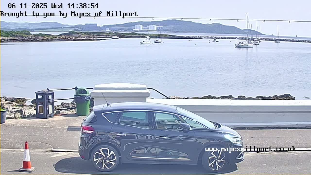 A dark-colored SUV is parked near a waterfront on a sunny day, with several boats visible in the calm water and a hilly background.