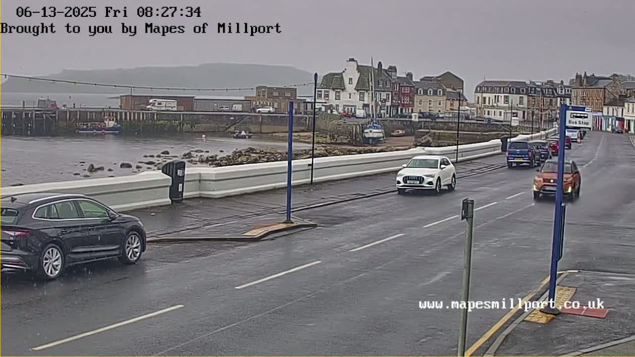 A wet road with several cars driving past a harbor, with a small town and hills visible in the background under an overcast sky.