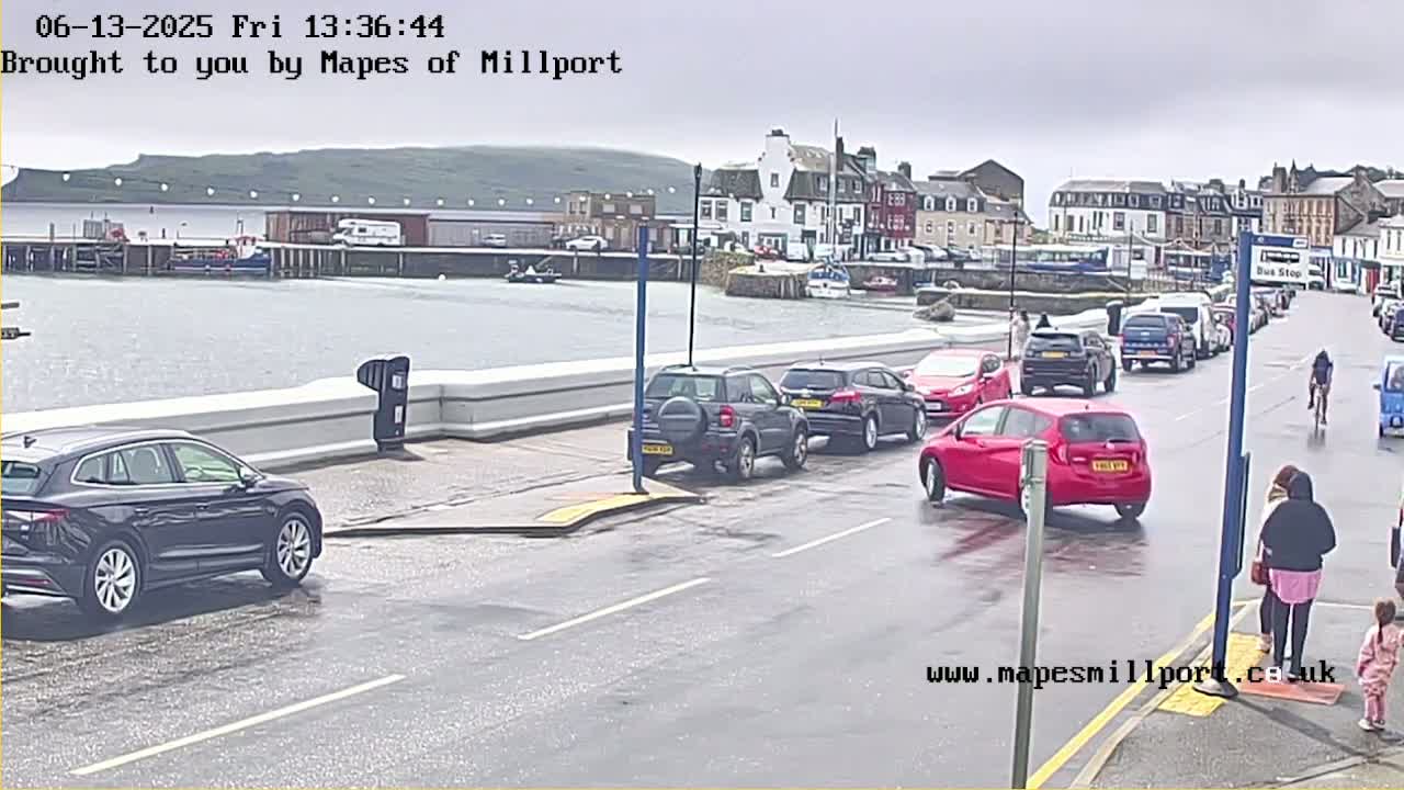 A wet street scene shows several cars parked and driving near a harbor, with a town and hills in the background under an overcast sky.