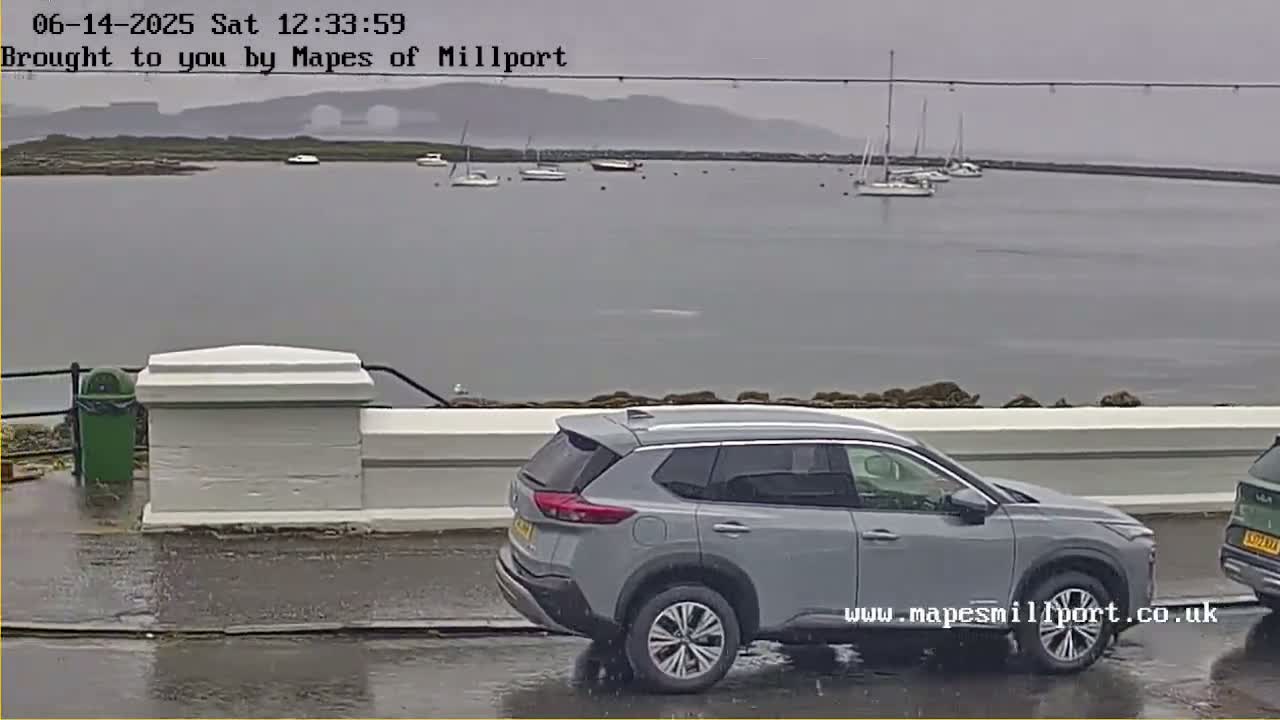 A silver SUV is parked on a wet street overlooking a calm body of water with several sailboats and a hazy mountain range in the background under an overcast sky.