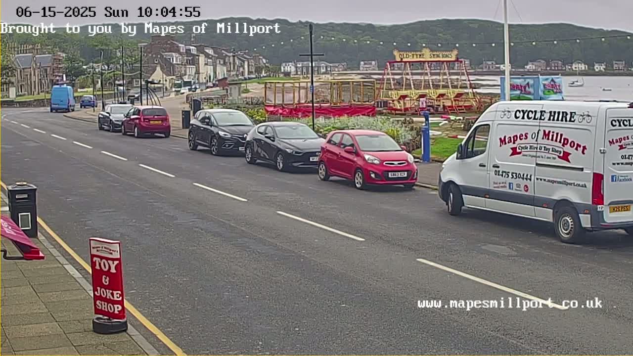 A street scene shows several cars parked along the side of a road, next to a seaside area with a carousel and a white van parked further down the road, on an overcast day.