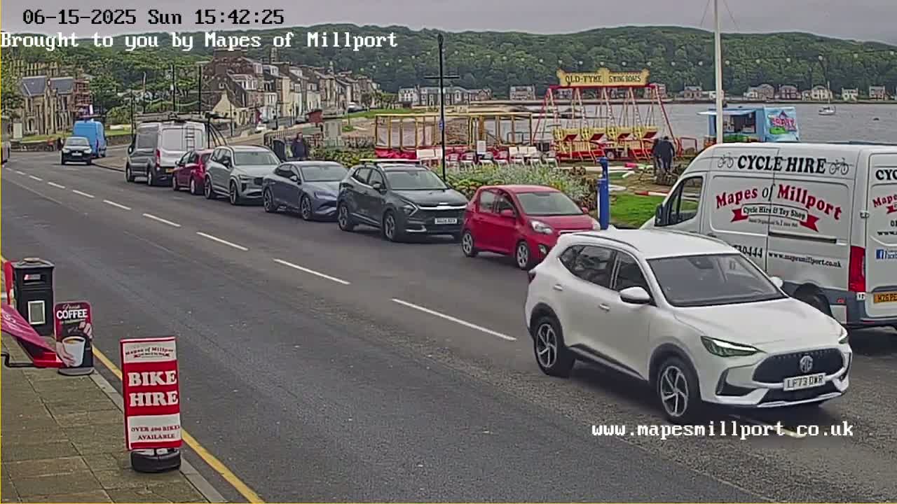 Several cars are parked along a street next to a waterfront, under an overcast sky.