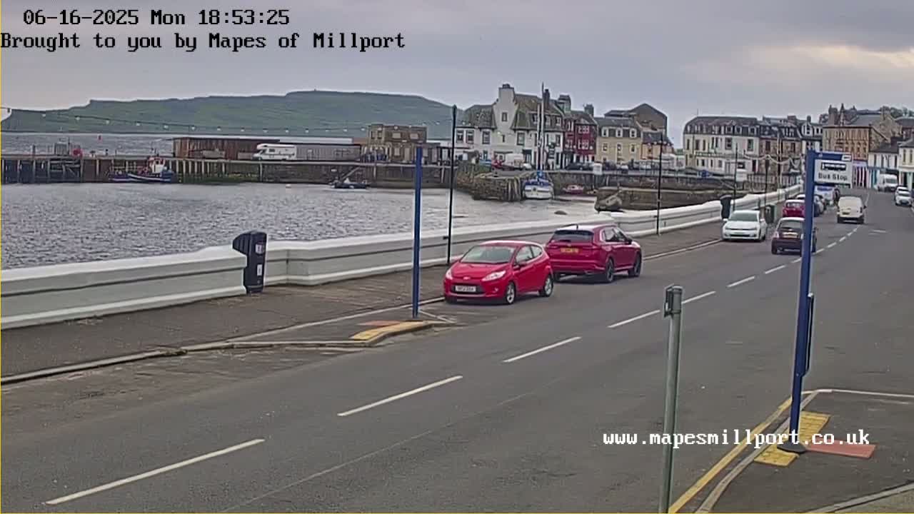 A seaside town is seen under overcast skies, with several cars driving along a road beside a harbor.