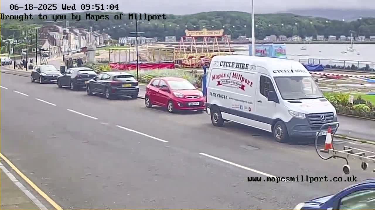 Several cars are parked along a street near a seaside amusement park on an overcast day.