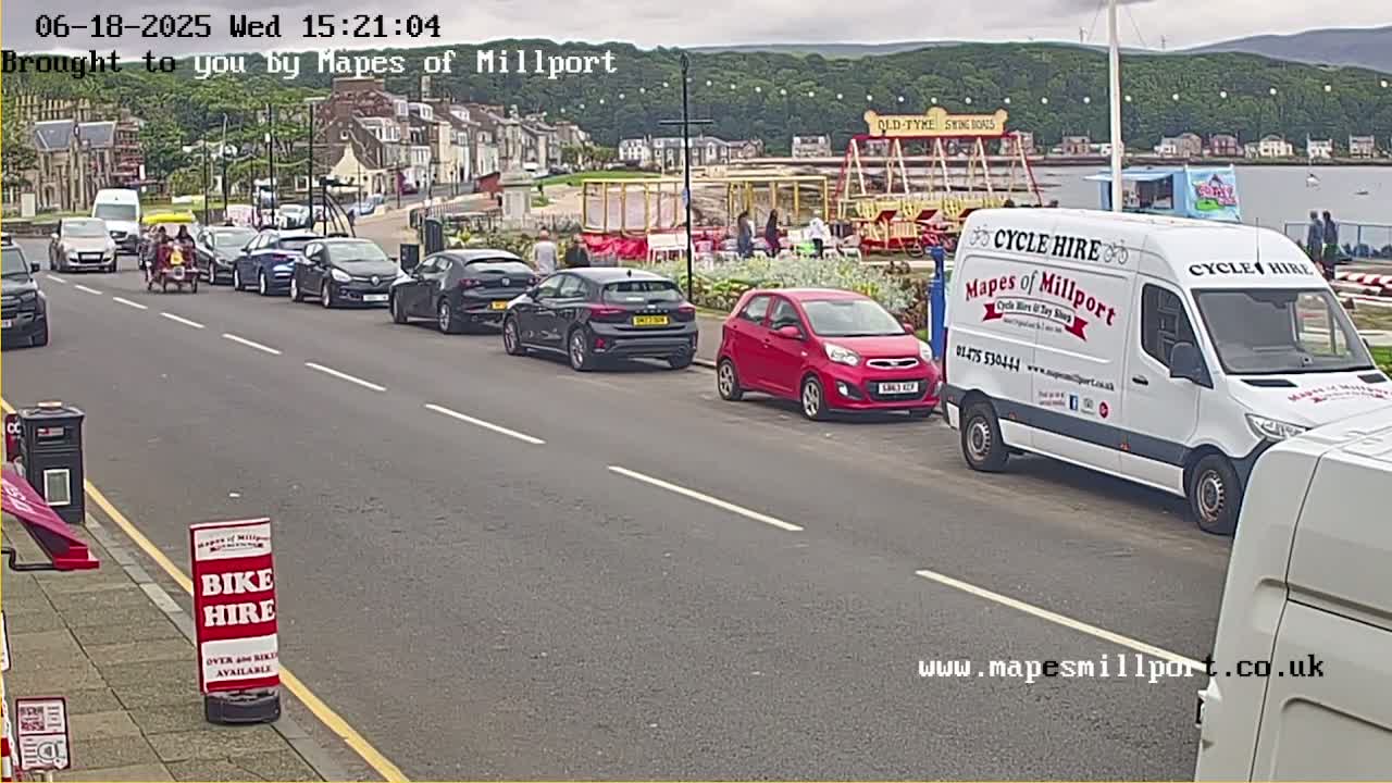 A street scene shows several cars parked along the side of a road, near a seaside town with a fairground ride visible in the background, under partly cloudy skies.