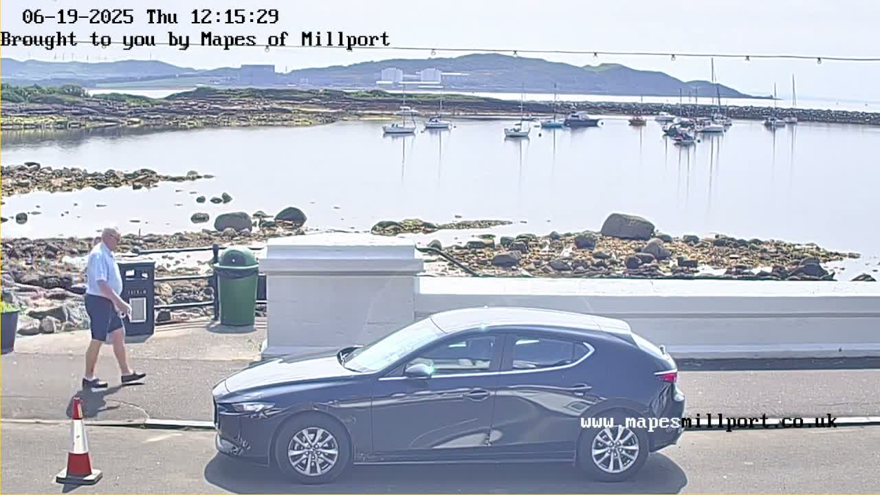 A dark-colored car is parked near a man walking on a sidewalk overlooking a calm body of water with several boats and a rocky shore under a sunny sky.