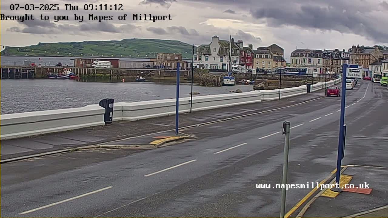 A harbor scene under cloudy skies shows a road with cars and a bus stop in the foreground, a low white wall separating the road from a harbor with boats, and buildings and a hill in the background.