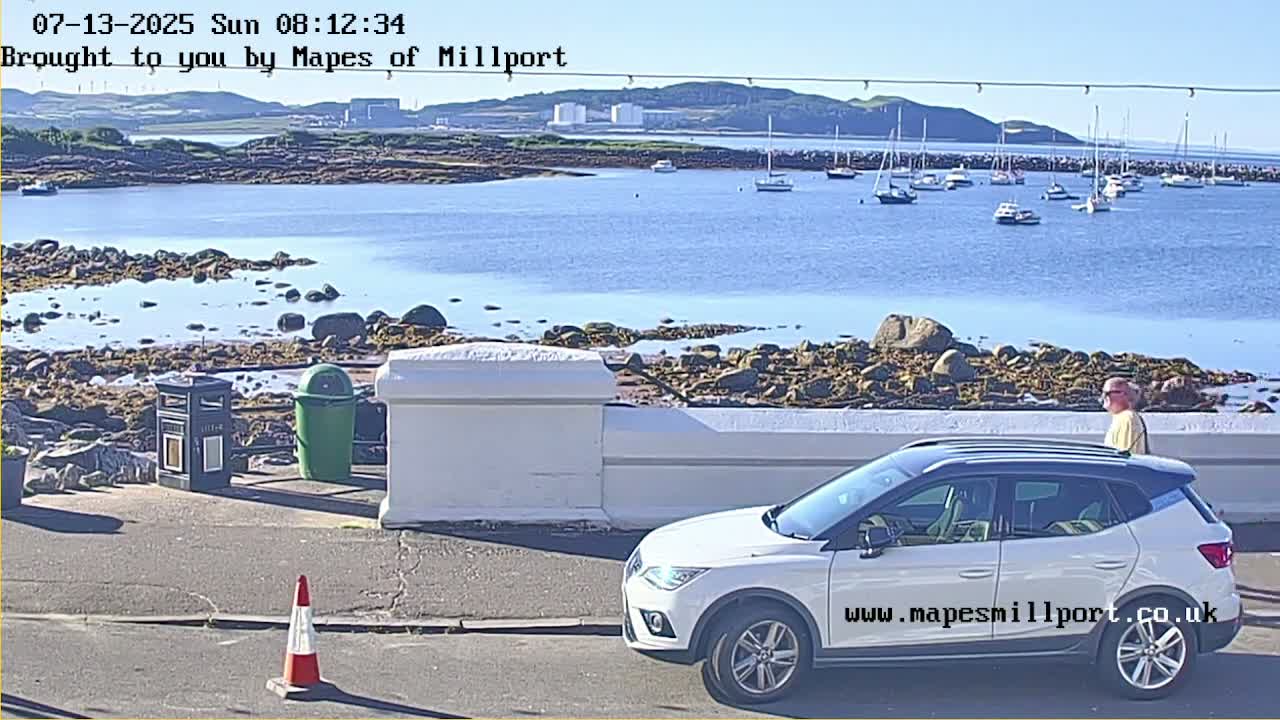 A sunny view of a calm harbor with numerous boats, a rocky shoreline, and a white SUV parked near a low wall, with a man walking nearby.