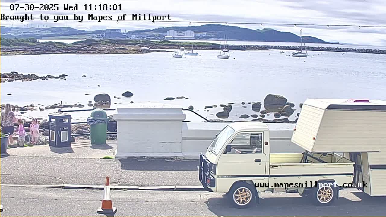 A sunny view of a calm harbor with numerous boats, a rocky shoreline, and a white SUV parked near a low wall, with a man walking nearby.