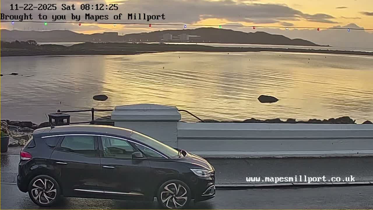 A dark car is parked beside a low wall, overlooking a calm body of water reflecting the golden light of a sunrise or sunset under a partly cloudy sky, with distant hills and a string of colorful lights hanging above the horizon.