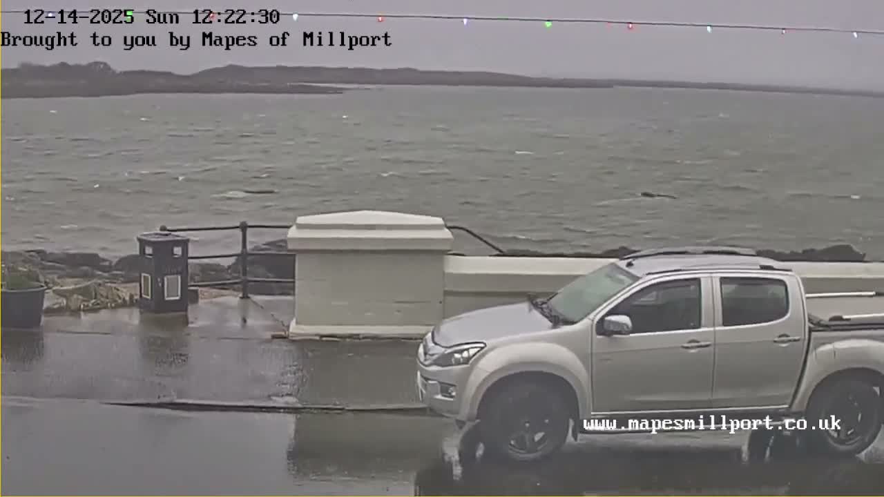 A dark car is parked beside a low wall, overlooking a calm body of water reflecting the golden light of a sunrise or sunset under a partly cloudy sky, with distant hills and a string of colorful lights hanging above the horizon.