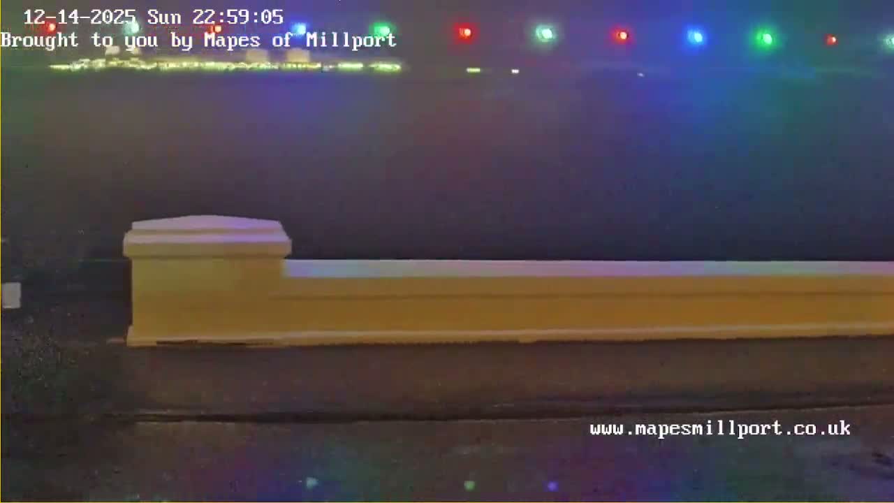 A dark car is parked beside a low wall, overlooking a calm body of water reflecting the golden light of a sunrise or sunset under a partly cloudy sky, with distant hills and a string of colorful lights hanging above the horizon.