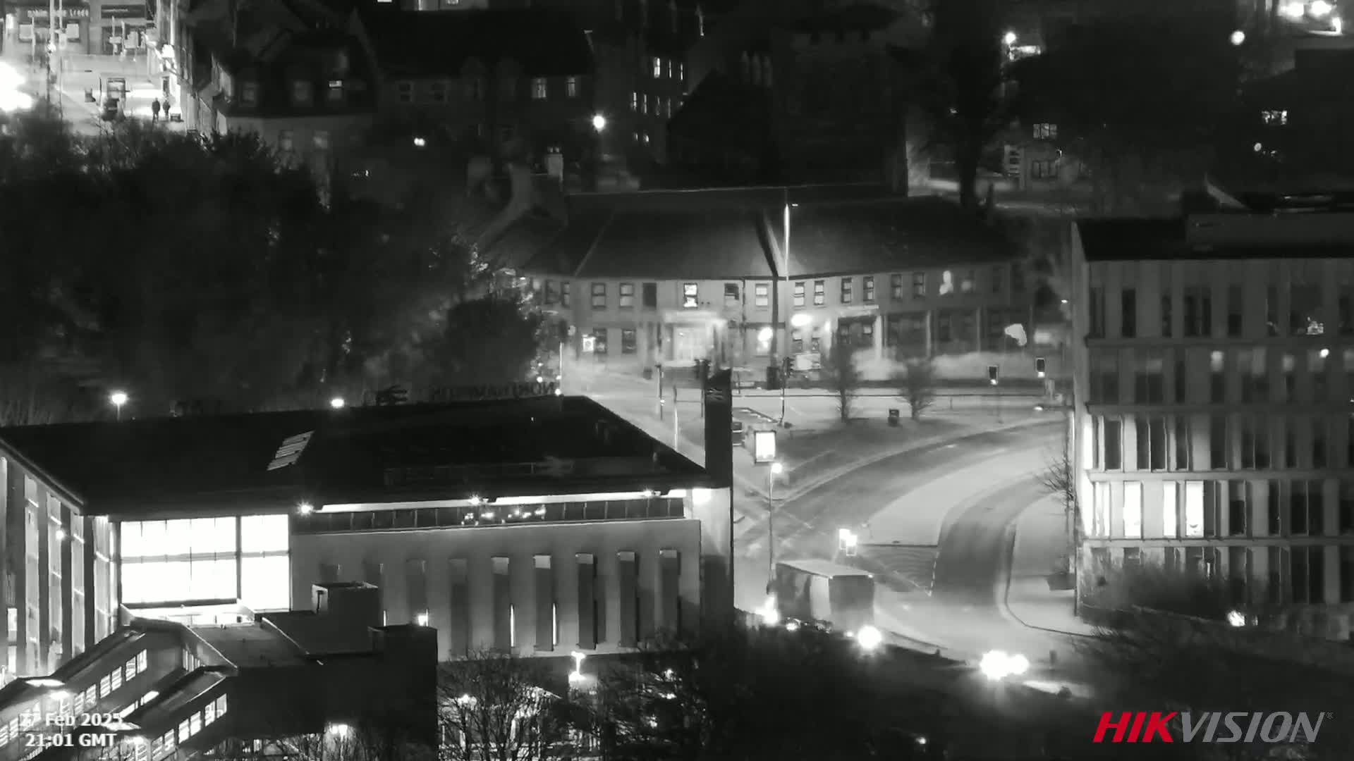 A nighttime aerial view of a snow-covered city street curving around buildings, with some vehicular traffic visible.