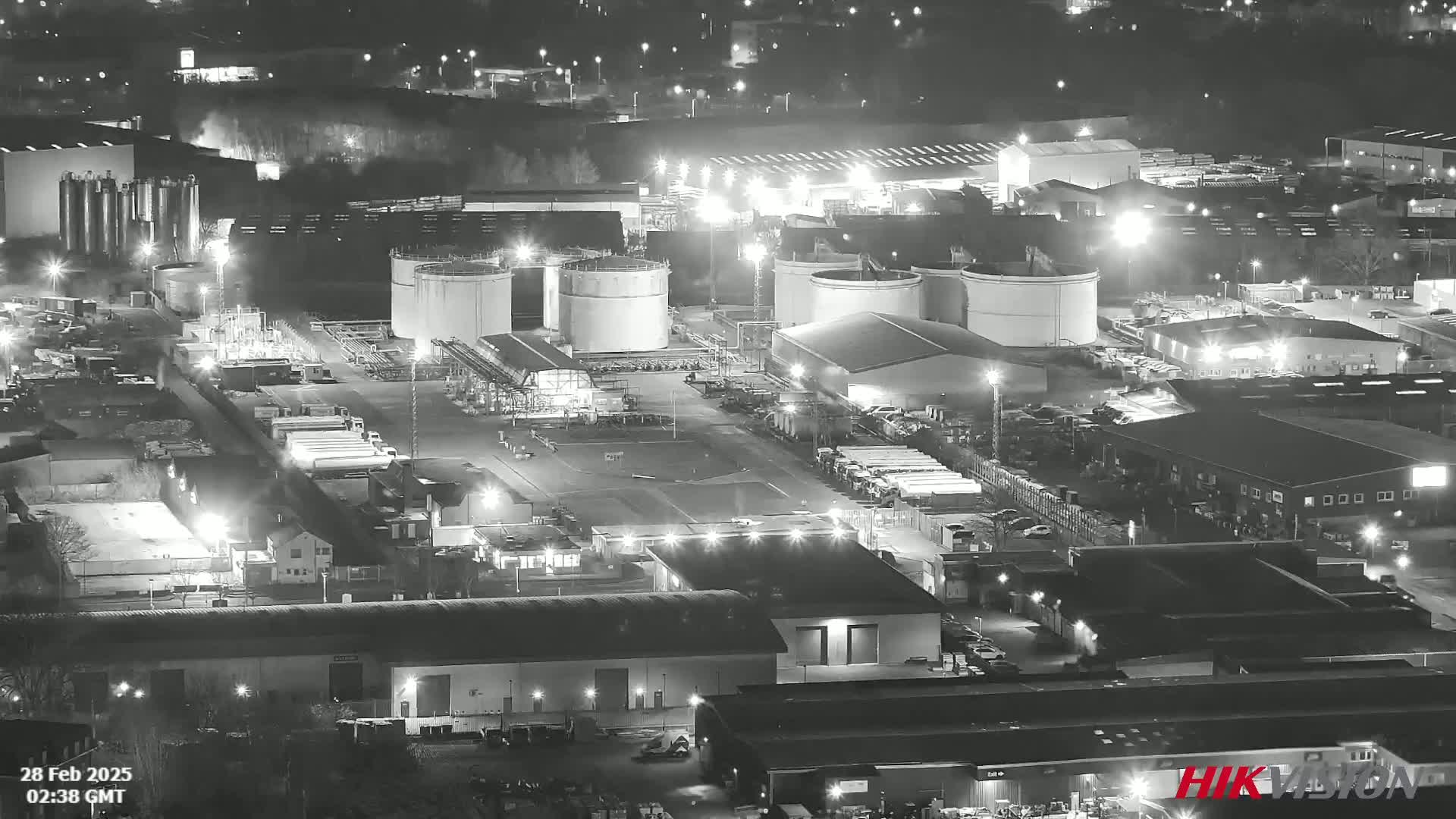 An illuminated industrial complex at night, featuring numerous large storage tanks and buildings, is shown in a black and white aerial view.