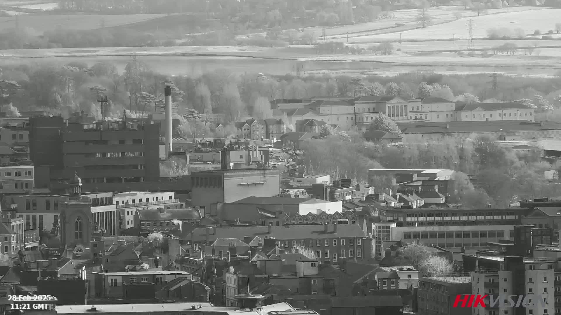 A grayscale aerial view of a city on a bright, hazy day shows numerous buildings, including industrial structures and larger, more formal buildings, surrounded by trees and fields.