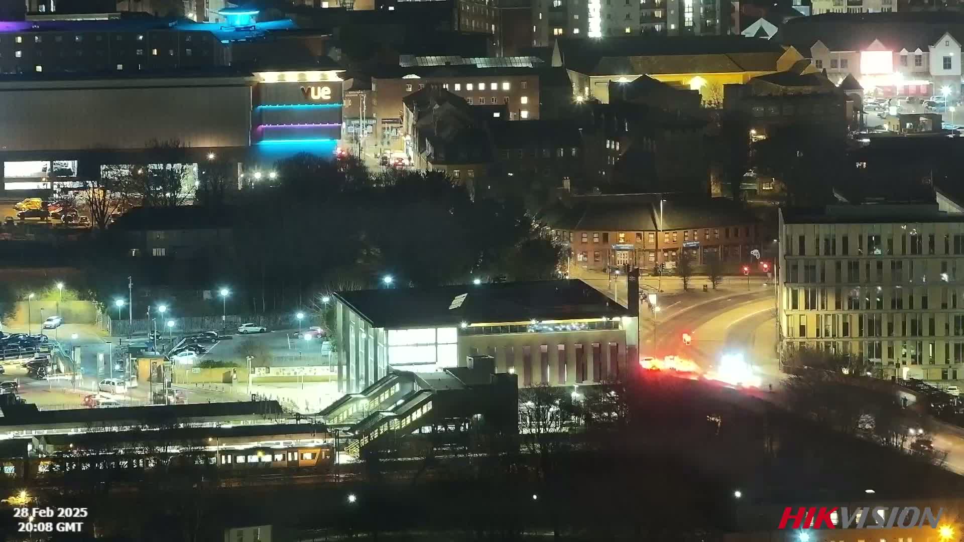 A nighttime aerial view of a city showing a train station, buildings, roads, and vehicles under clear skies.