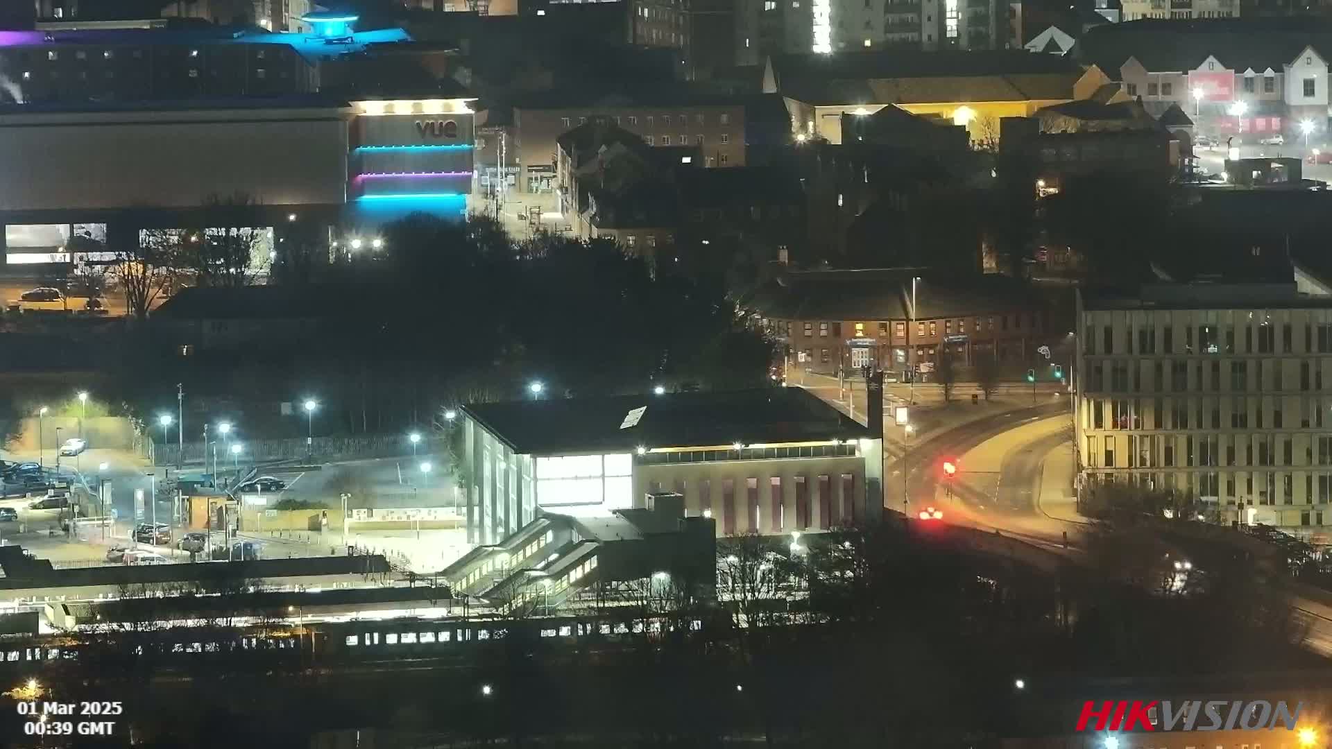 A nighttime aerial view of a city featuring illuminated buildings, a train station, and roads with minimal traffic.