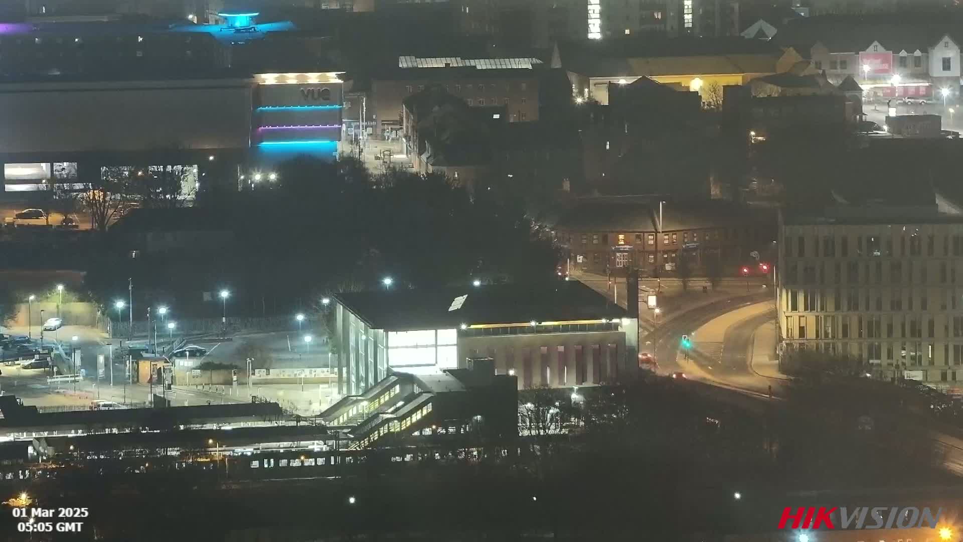 A nighttime aerial view of a city showing a train station, buildings, roads, and sparsely-lit streets under a hazy sky.
