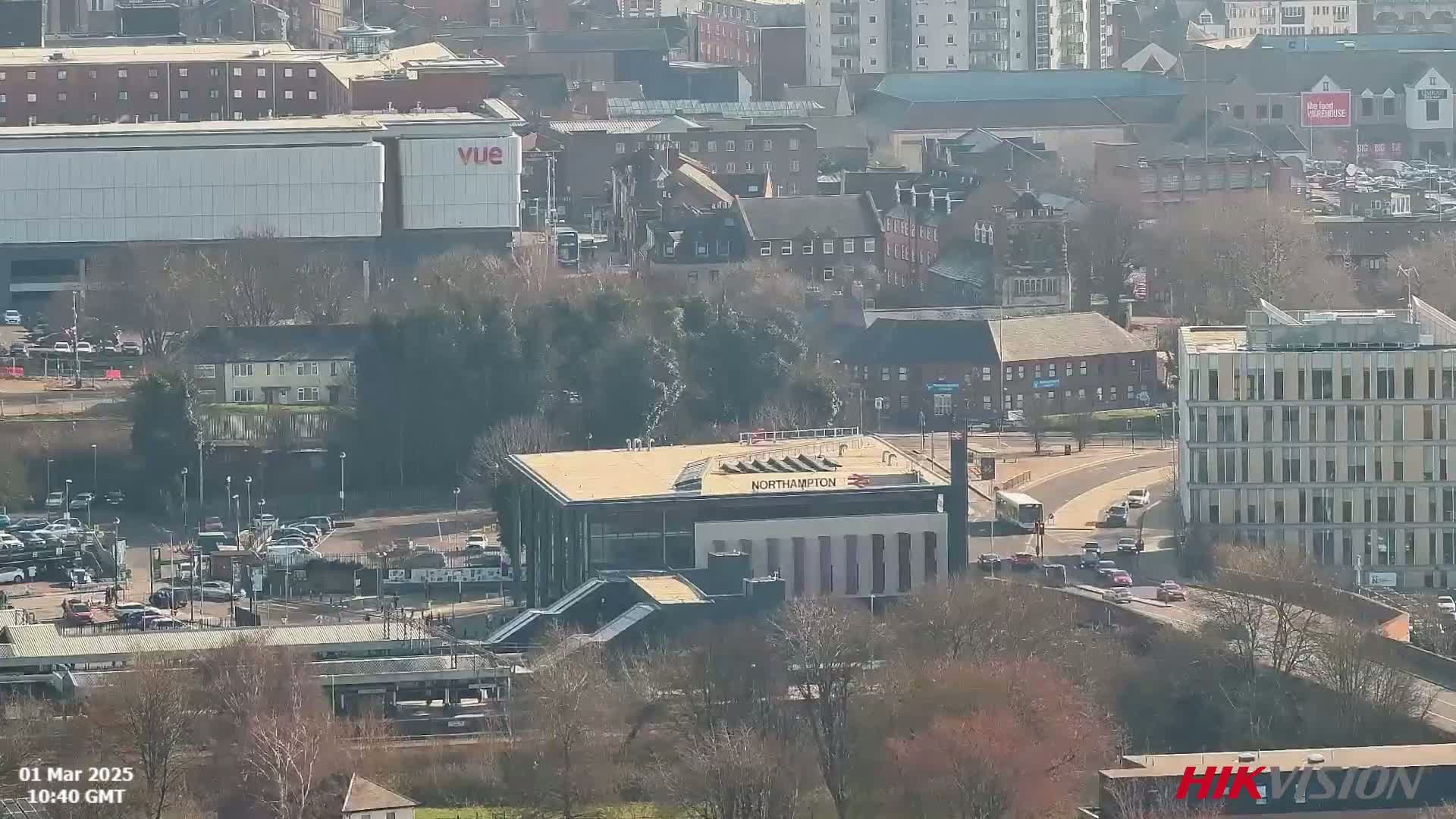 An aerial view shows a city scene on a sunny day, featuring a train station, buildings, roads with vehicles, and surrounding trees.