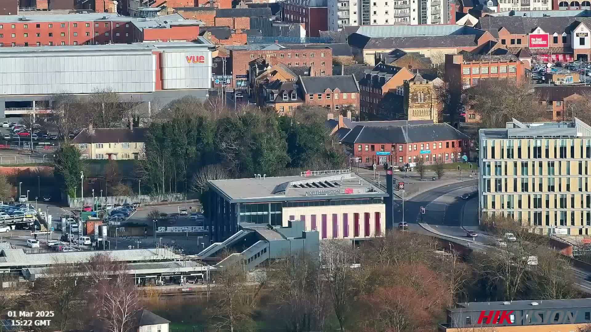 An aerial view shows a city on a sunny day with a modern train station and surrounding buildings, parking lots, and trees.