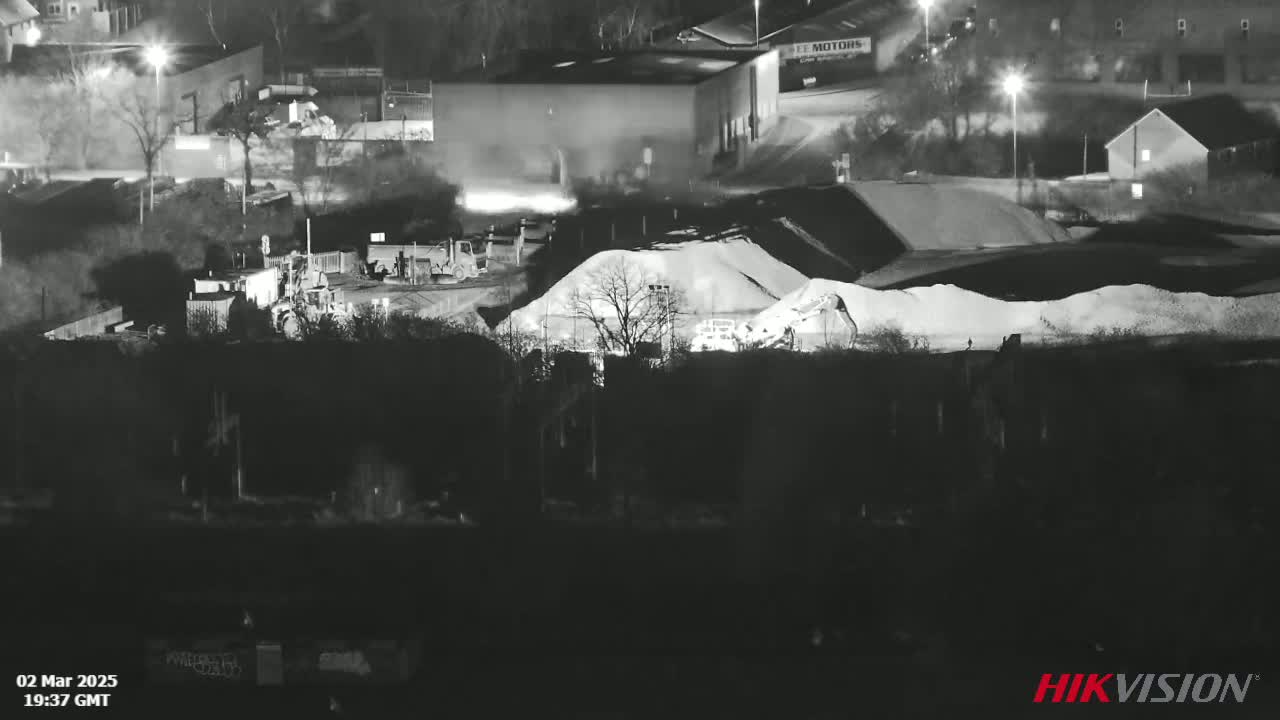 A nighttime, low-light view of an industrial site with piles of aggregate material, several pieces of heavy machinery, and surrounding buildings.