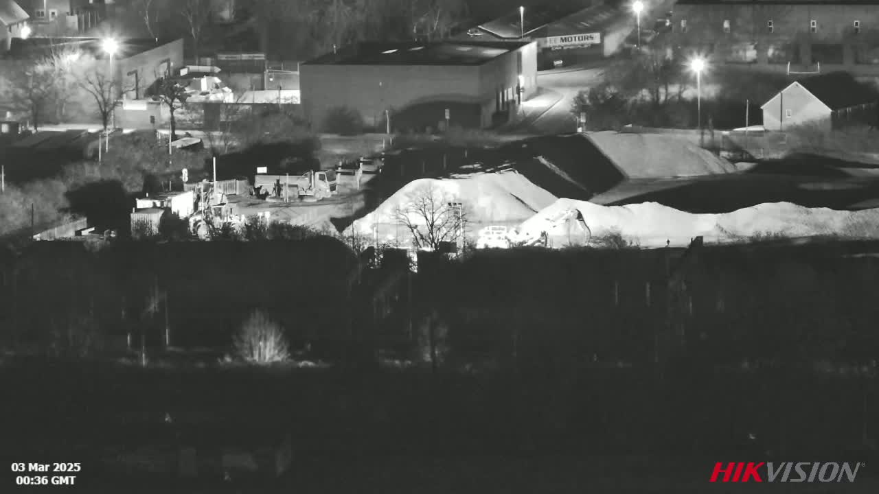 A nighttime, black and white view of heavy machinery and piles of aggregate material at an industrial site.