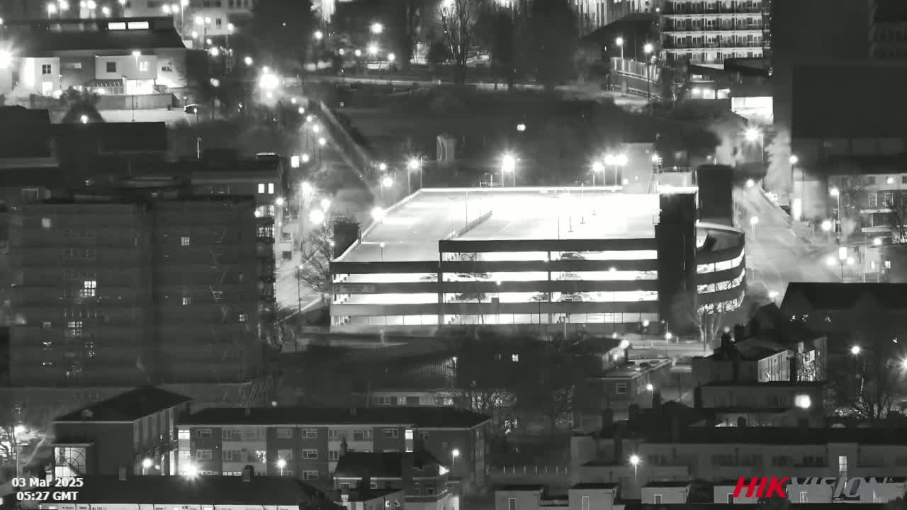 A nighttime, black and white aerial view shows a multi-level parking garage brightly lit amongst numerous buildings and streets with light snow or rain falling.