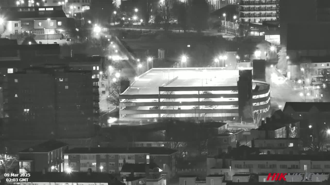 A nighttime, high-angle, black and white view of a city with a multi-level parking garage illuminated, and light mist or fog visible in the air.