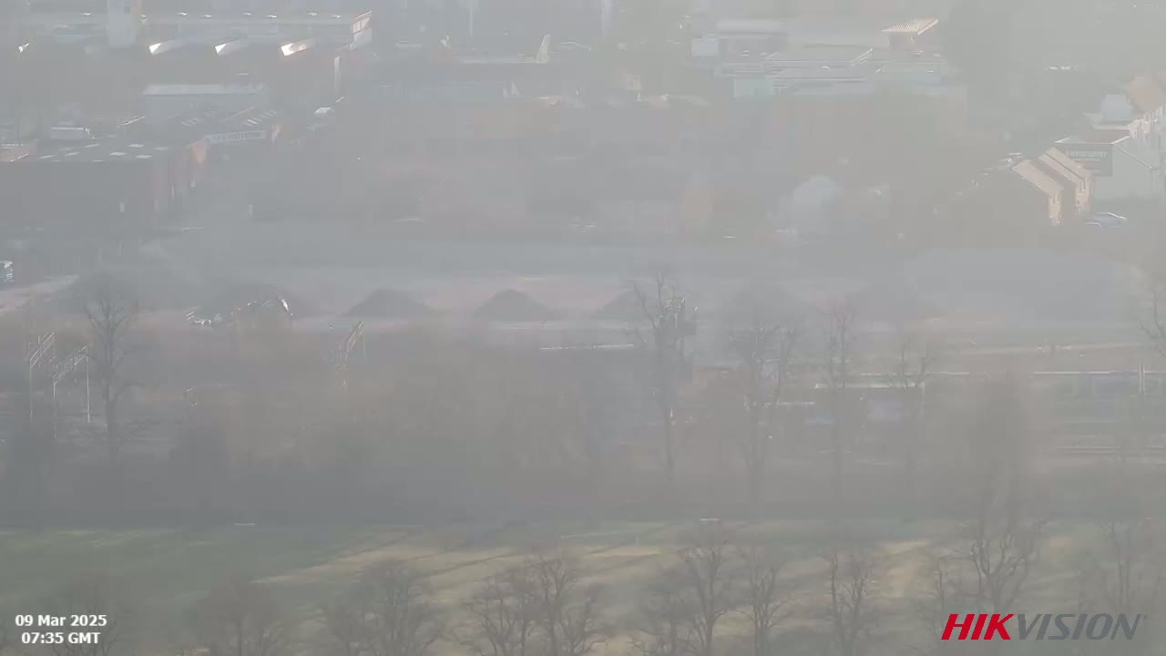 A hazy, foggy view of a low-lying area with buildings and bare trees, possibly a field or industrial area.