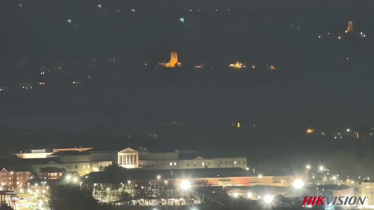 A nighttime long shot shows a cityscape with illuminated buildings, including a large, rectangular structure in the foreground and smaller lights scattered across a hill in the background under a dark sky.