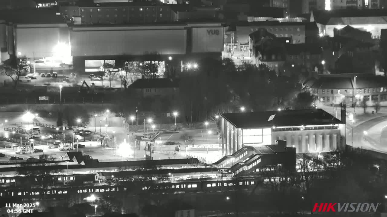 A nighttime, high-angle, black and white view of a train station and surrounding buildings, parking lots, and trees under clear skies.
