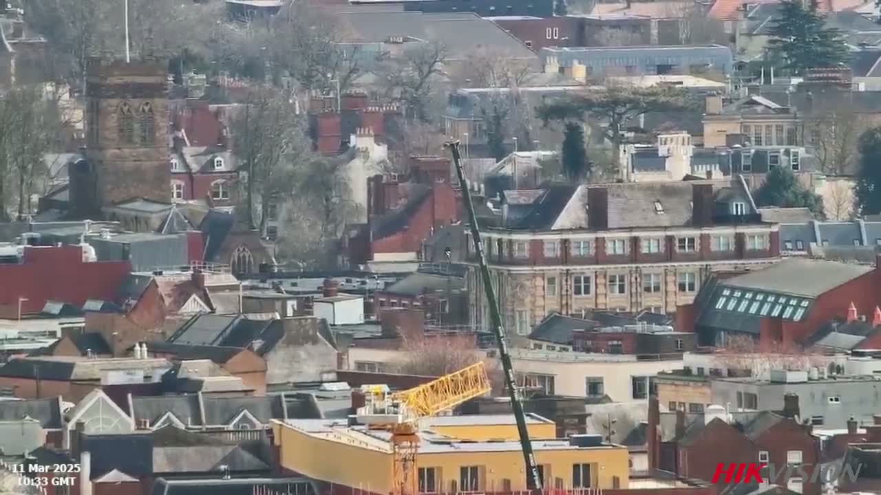 A dense collection of brick buildings, some with visible church steeples, are seen from a high vantage point on a mostly sunny day, with a yellow crane partially visible in the foreground.