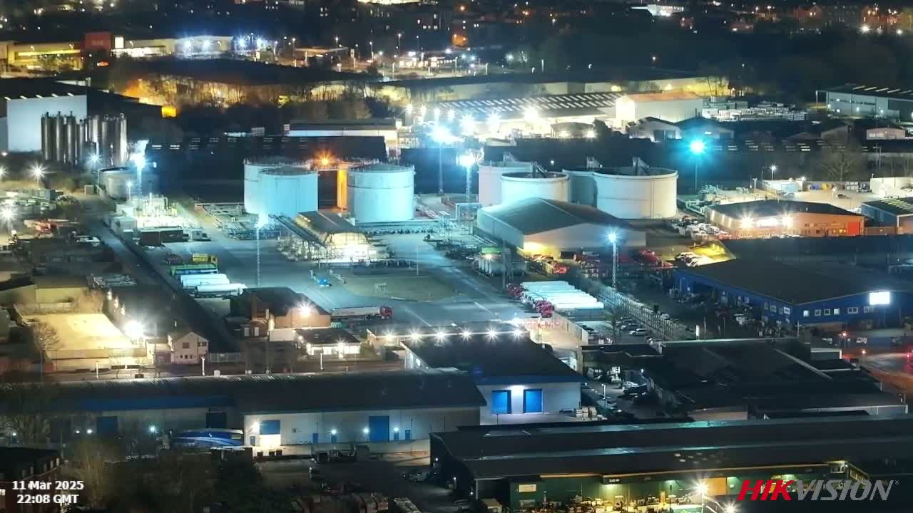 An industrial complex at night, brightly lit, with numerous large storage tanks and buildings, surrounded by trucks and other vehicles.