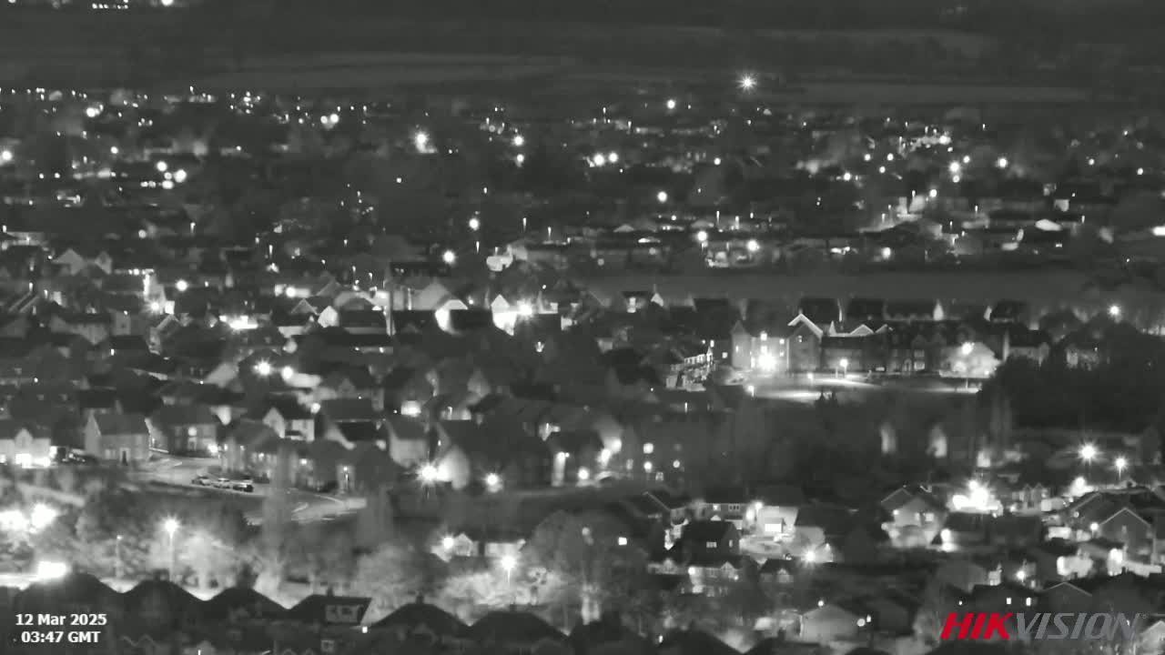 A nighttime aerial view shows a residential area with numerous houses illuminated by streetlights and house lights under a clear, dark sky.
