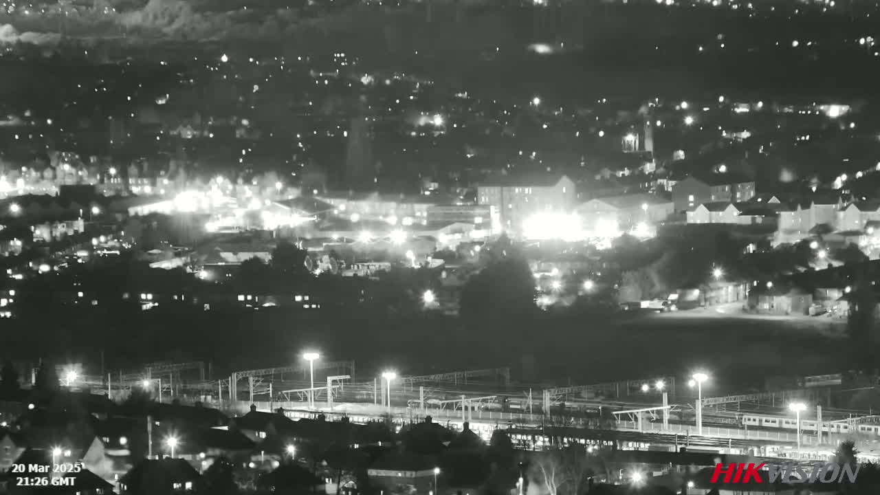 A nighttime, black and white long shot shows a sprawling city with illuminated buildings and train tracks, under a hazy sky.