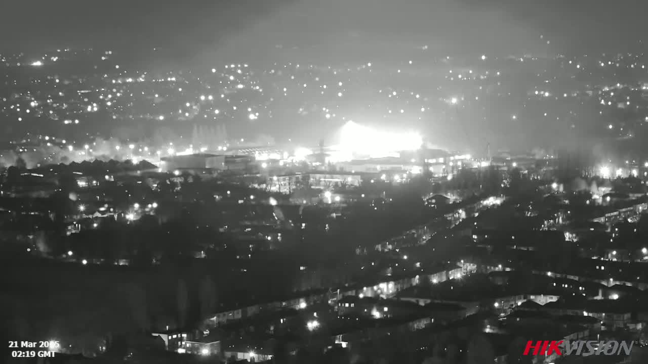 A nighttime, long-range grayscale view of a city with numerous lights, showing a large bright area possibly a fire or explosion in the mid-ground.