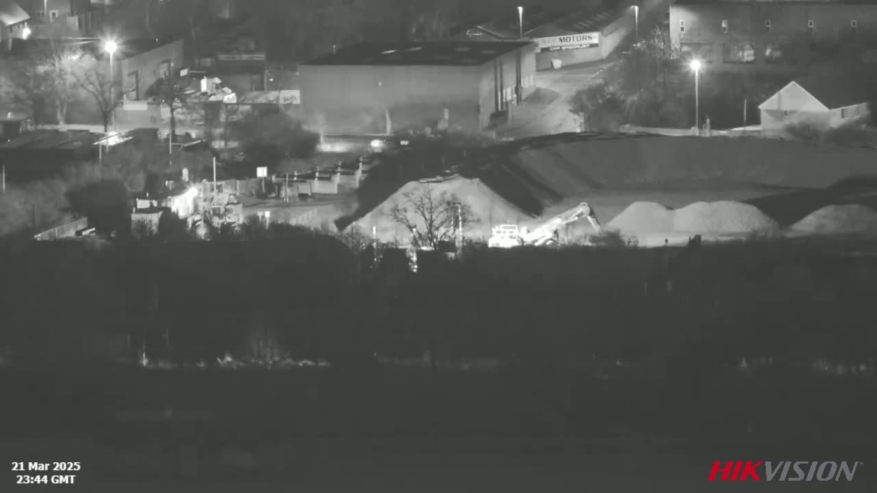 A nighttime, low-light image shows heavy machinery working near large piles of aggregate material in an industrial area.
