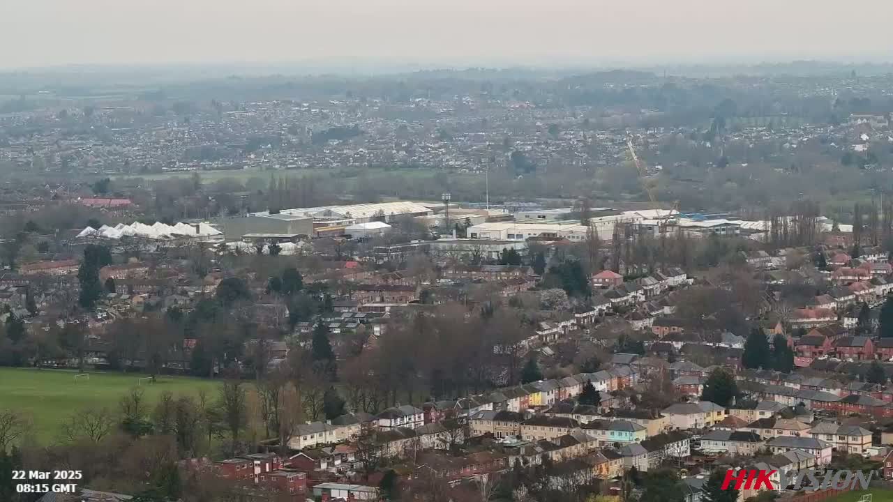 A sprawling residential area with a large industrial complex and sports field is visible on an overcast day.