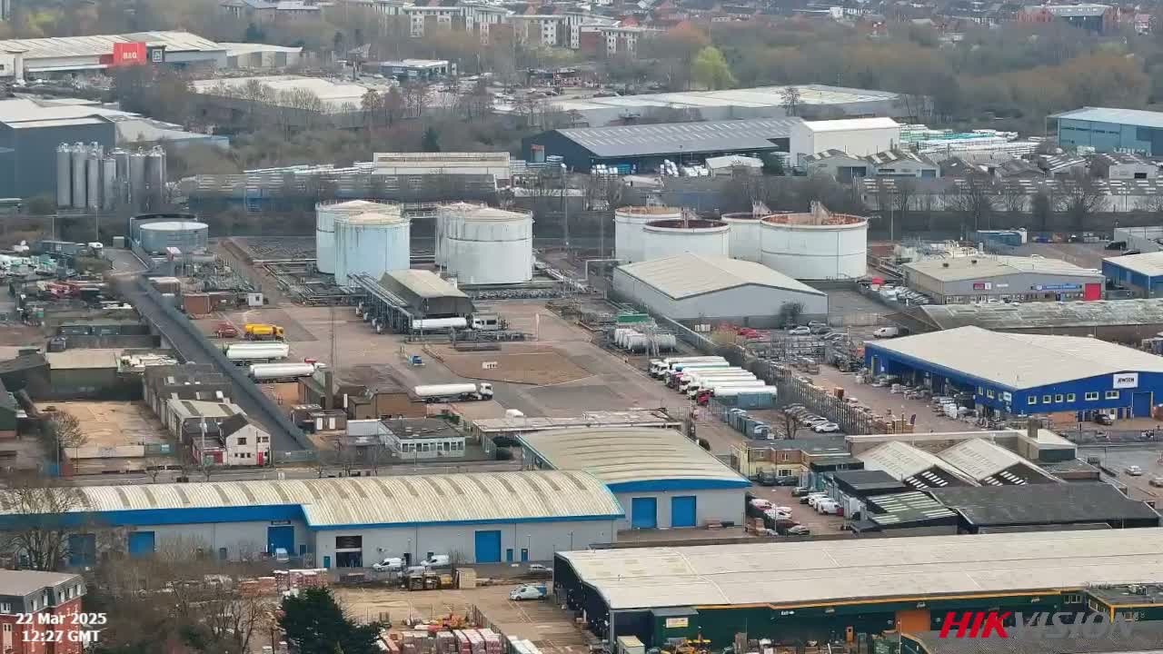 An aerial view of an industrial area with numerous buildings, large storage tanks, and many parked vehicles, on an overcast day.