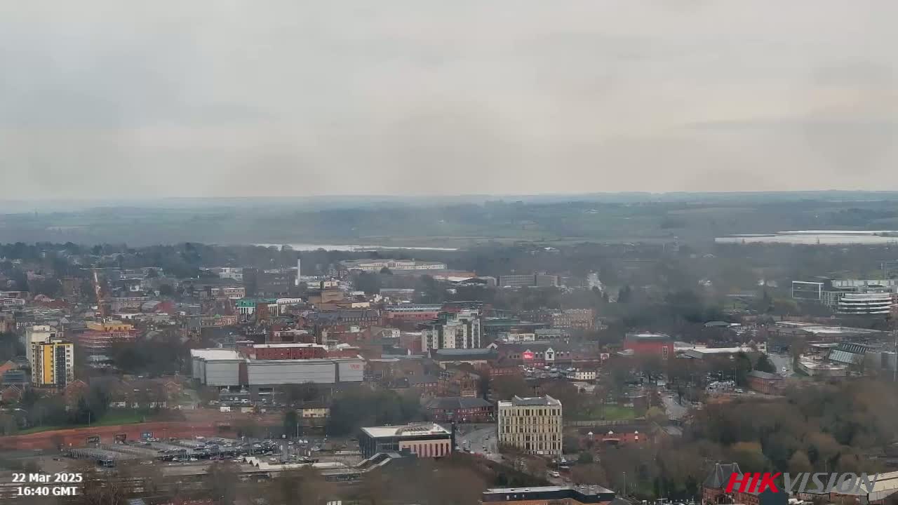 A hazy, overcast view shows a sprawling city with numerous buildings and trees, stretching to a river visible in the distance.