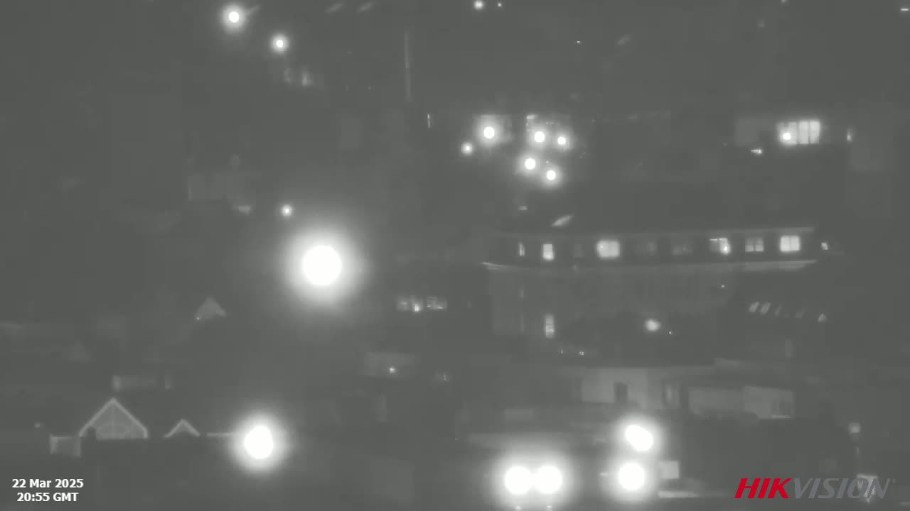 A nighttime, low-light grayscale image shows a street scene with buildings, vehicles, and streetlights.