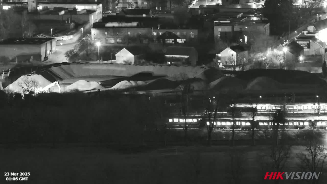 A nighttime, black and white aerial view shows a train passing by a large pile of sand or gravel next to a residential area.