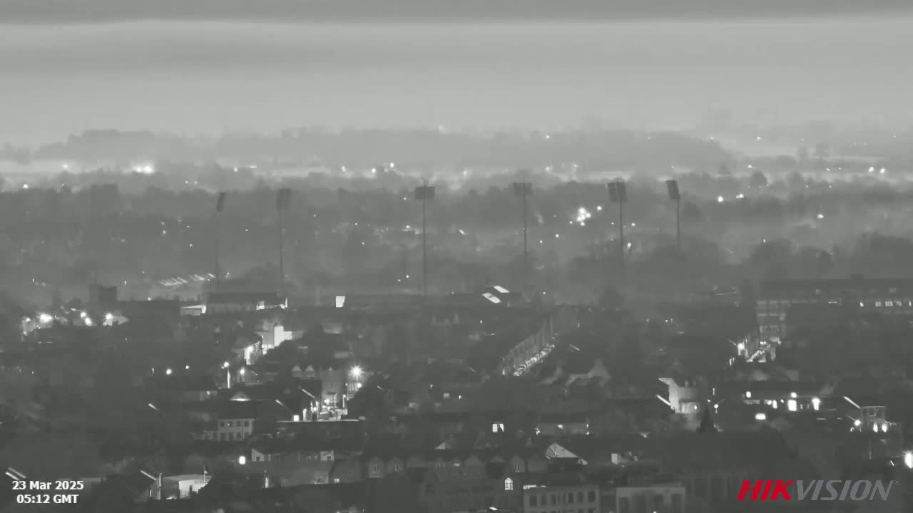 A foggy, nighttime view of a city with several tall lights in the middle ground, showing a residential area with scattered lights.