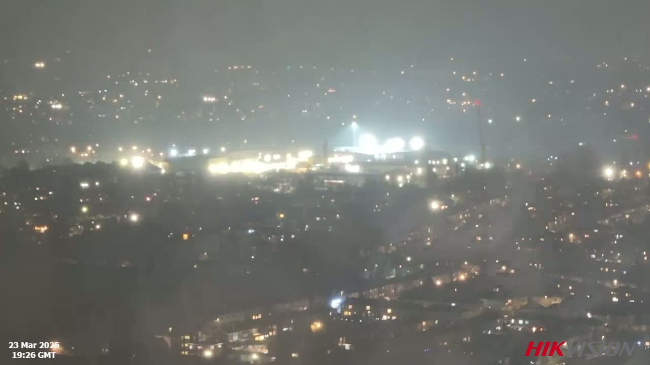 A nighttime view of a city shrouded in fog, with brightly lit buildings and a stadium visible in the distance.