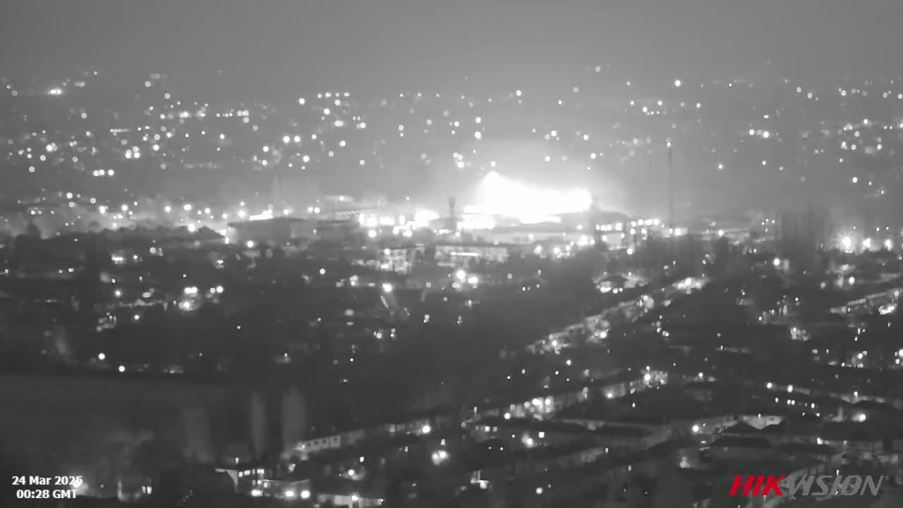 A nighttime, long-range grayscale view of a city with many lights, showing a brightly lit area in the mid-ground that appears to be a stadium or large building.