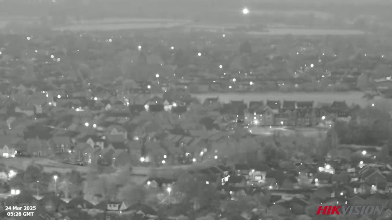 A grayscale night vision image shows a sprawling residential area with scattered lights under a hazy sky.